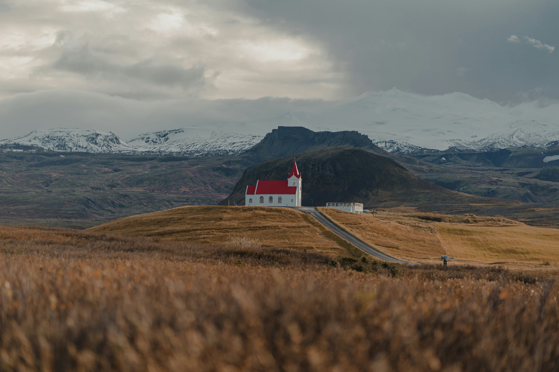 A white church with a red roof on a grassy hill, with a road running up to it and snow-covered mountains behind