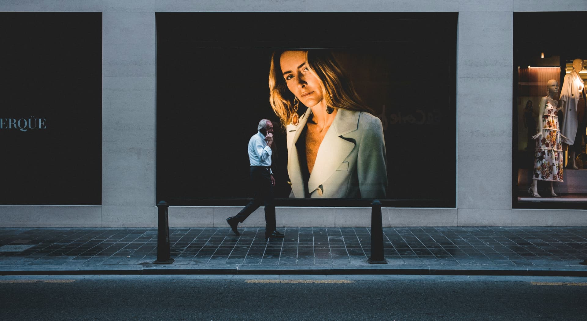 A man walks along a city sidewalk on a phone call, passing a storefront advertisement far taller than he is