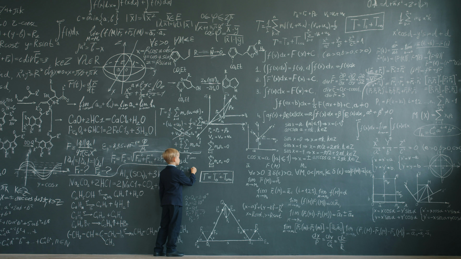 A boy in a school uniform writes on a blackboard covered floor to ceiling in equations
